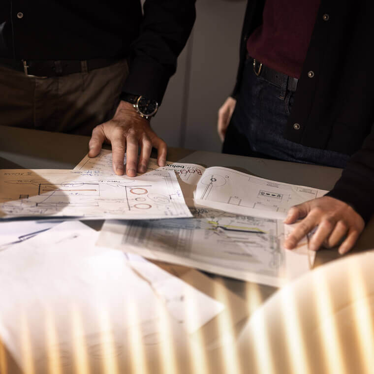 Zwei Personen betrachten technische Zeichnungen auf einem Tisch; daneben liegen helle, ringförmige Leuchtenteile.