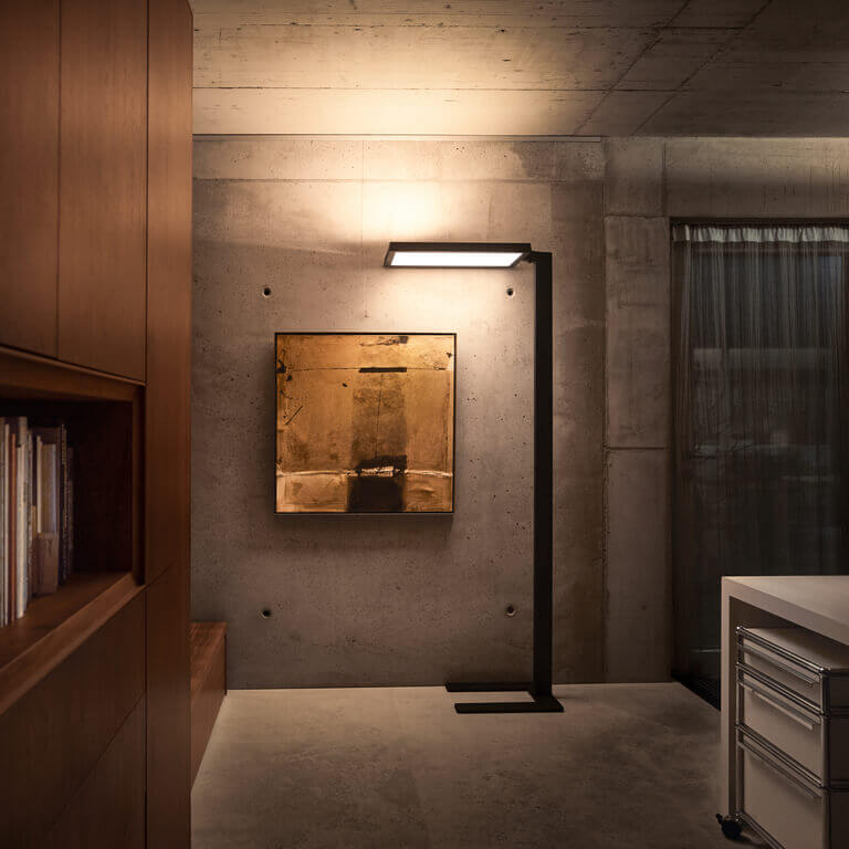 Schwarze Belux-Stehleuchte „Bob“ mit rechteckigem Leuchtpanel vor Sichtbetonwand, neben Bücherregal und Bild.