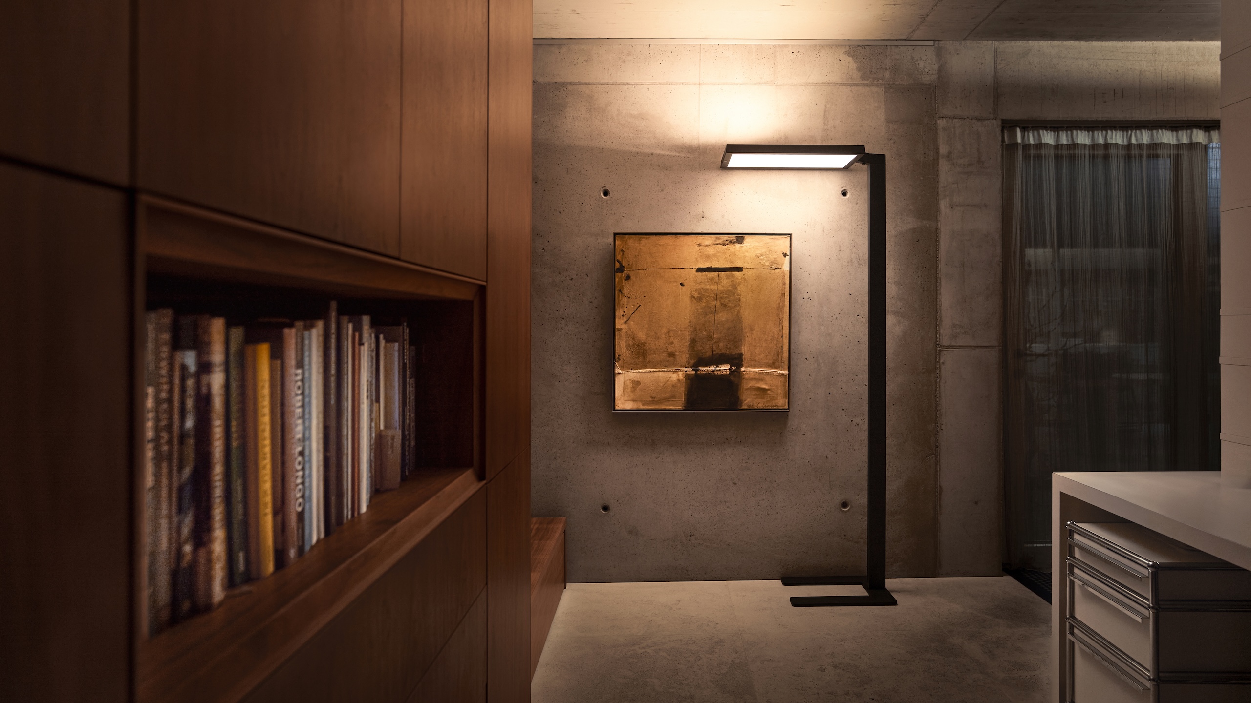 Schwarze Belux-Stehleuchte „Bob“ mit rechteckigem Leuchtpanel vor Sichtbetonwand, neben Bücherregal und Bild.