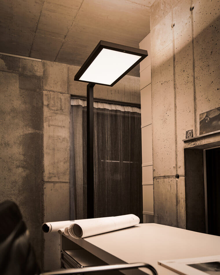 Belux-Stehleuchte „Bob“ mit großem, rechteckigem Leuchtpanel neben Tisch mit gerollten Plänen.