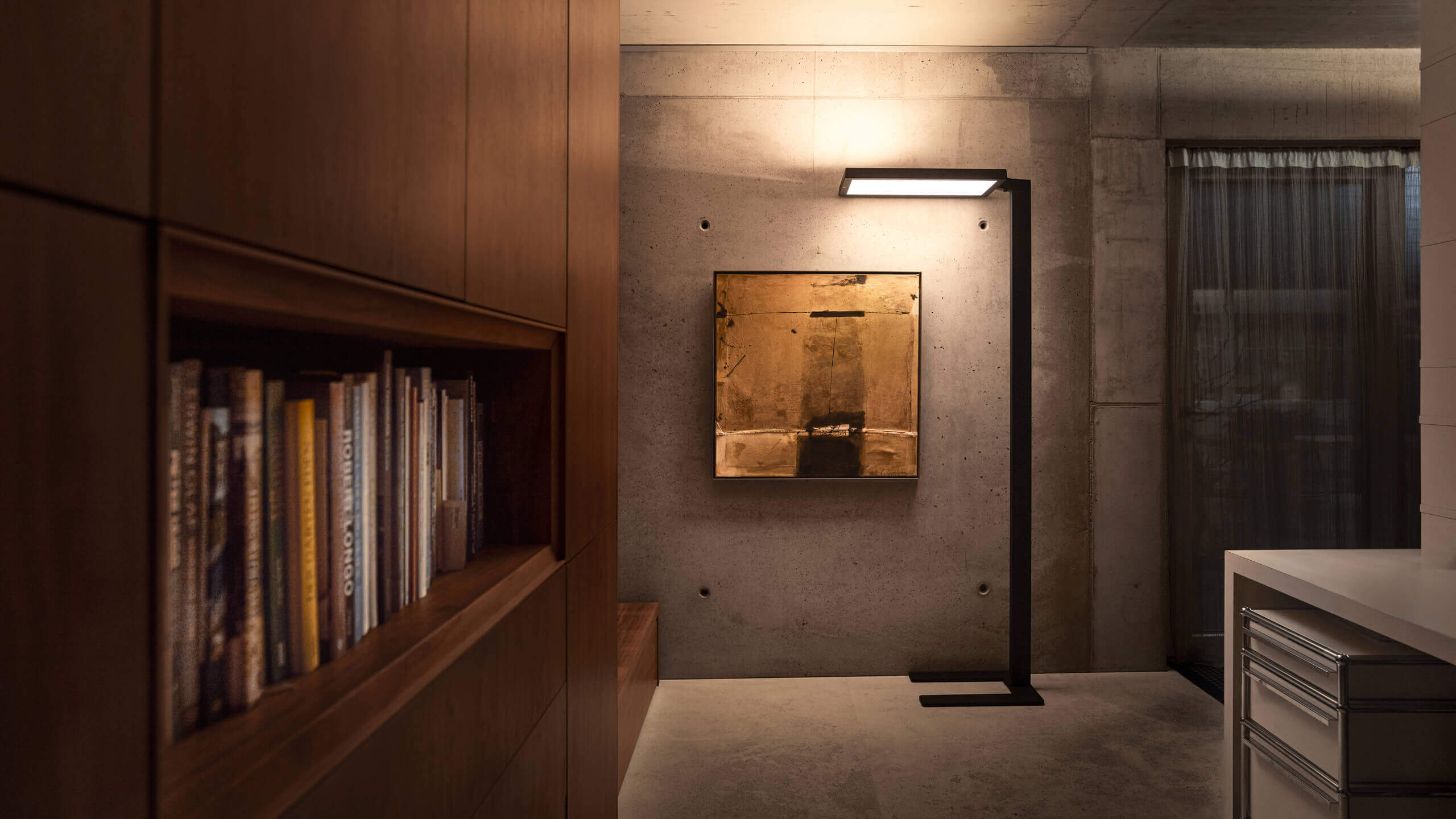 Schwarze Belux-Stehleuchte „Bob“ mit rechteckigem Leuchtpanel vor Sichtbetonwand, neben Bücherregal und Bild.