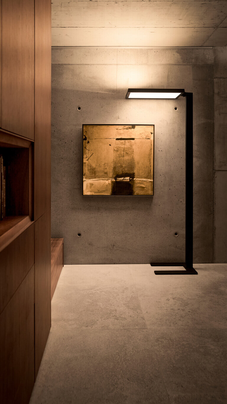 Schwarze Belux-Stehleuchte „Bob“ mit rechteckigem Leuchtpanel vor Sichtbetonwand, neben Bücherregal und Bild.