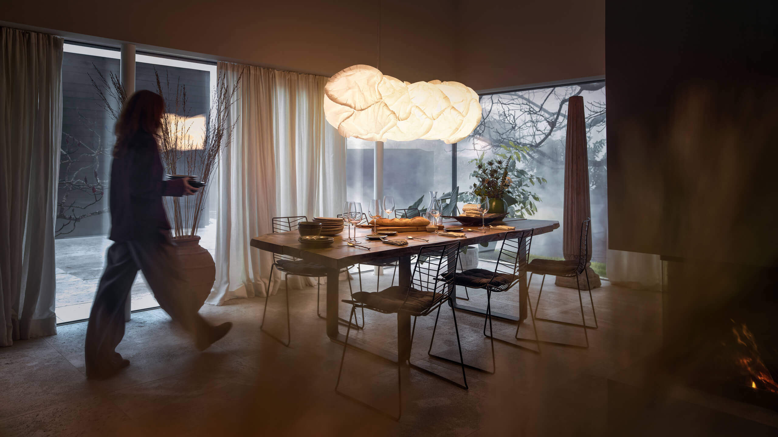 Esszimmer mit gedecktem Tisch und großen Fenstern; darüber hängt die leuchtende Pendelleuchte Belux „Cloud“, links läuft eine Person vorbei.