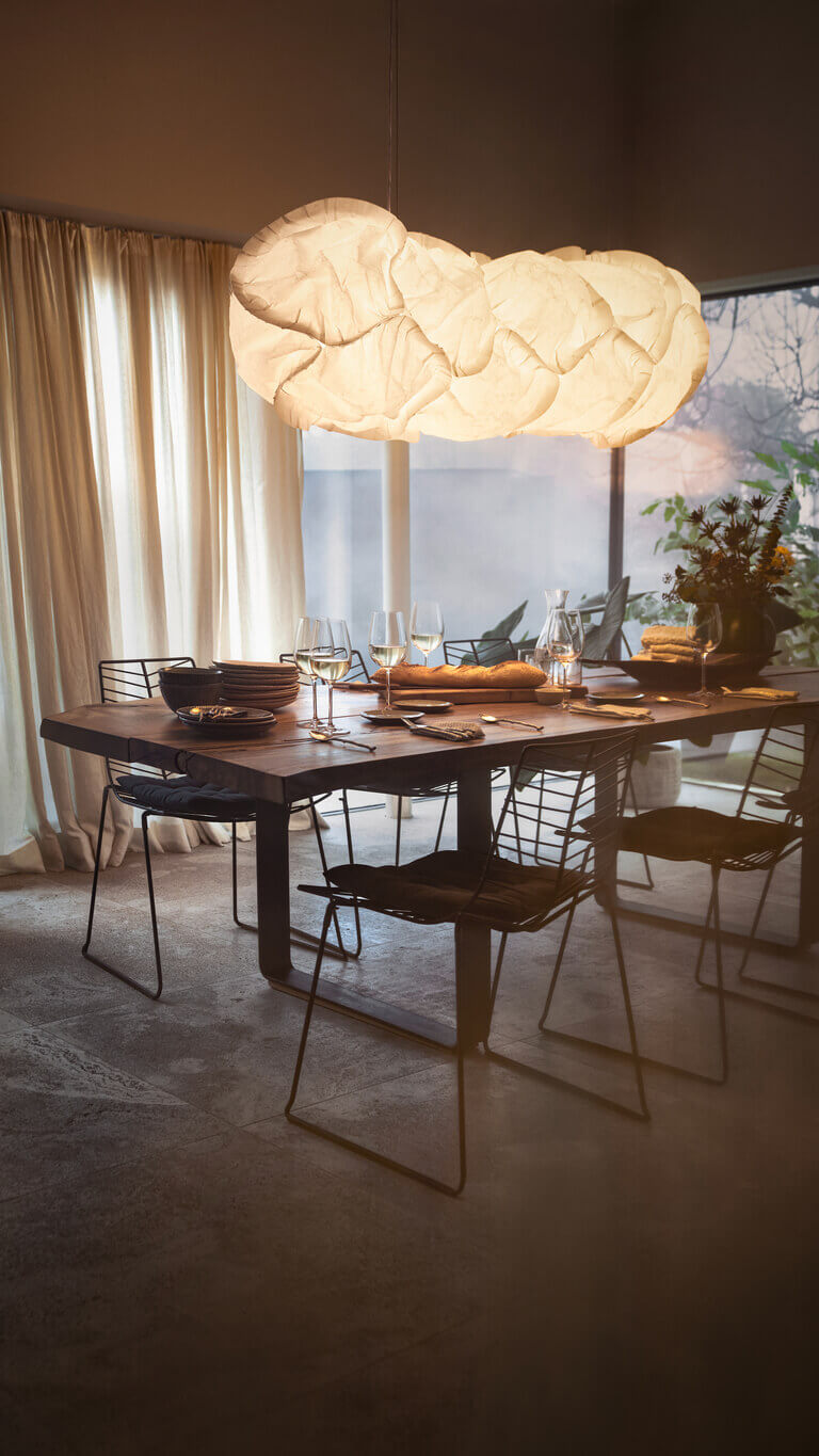 Esszimmer mit gedecktem Tisch und großen Fenstern; darüber hängt die leuchtende Pendelleuchte Belux „Cloud“, links läuft eine Person vorbei.