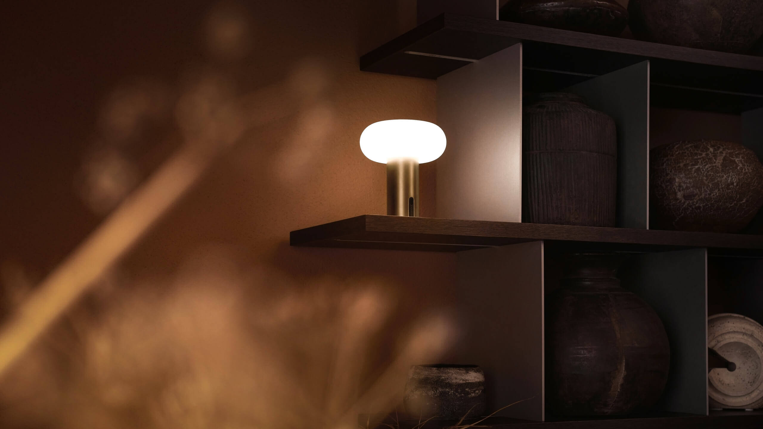 Belux „Oto Portable“ leuchtet in einer Regalnische; dunkle Keramikobjekte stehen daneben.