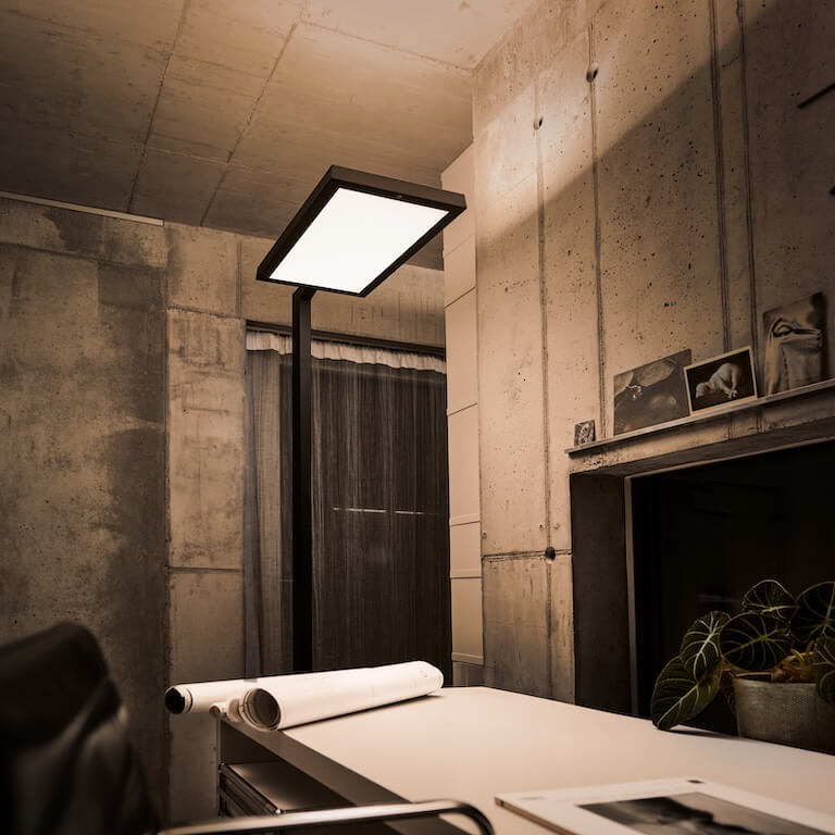 Belux-Stehleuchte „Bob“ mit großem, rechteckigem Leuchtpanel neben Tisch mit gerollten Plänen.