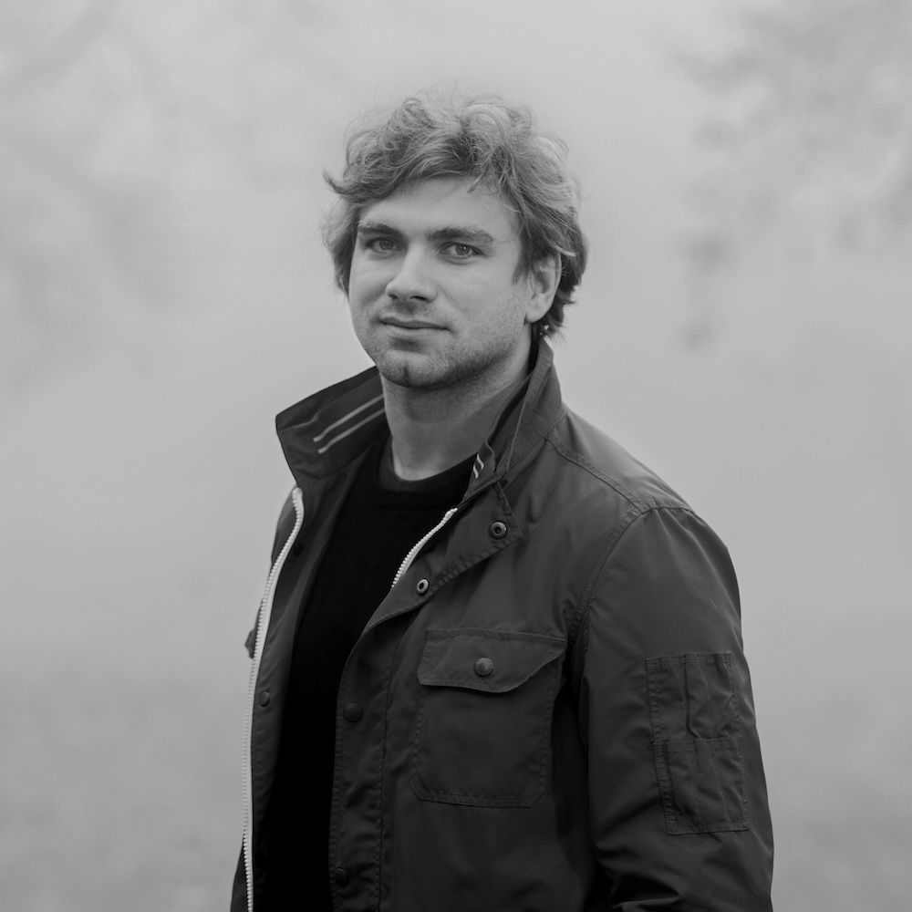 Der Designer Karel-Matejka mit lockigem Haar in einem Feld, trägt eine Jacke und schaut in die Kamera. Schwarz-weiß Portrait Foto. Er ist der Designer des Lampenmodells Moiré der Firma Belux.