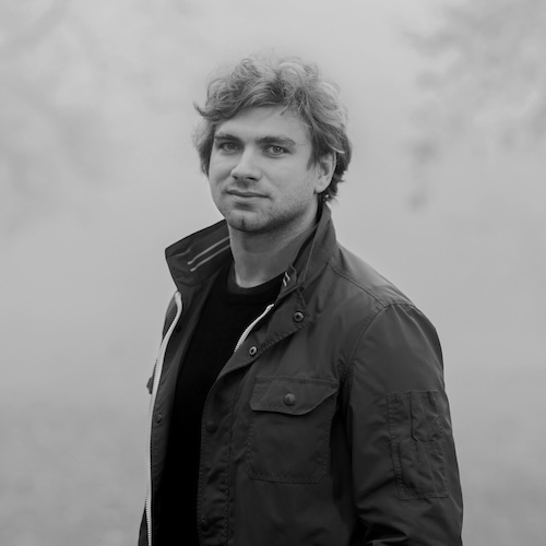 Designer Karel Matejka, with curly hair, stands in a field wearing a jacket and looking at the camera. Black and white portrait photograph. He is the designer of the Moiré lamp model for the Belux company.