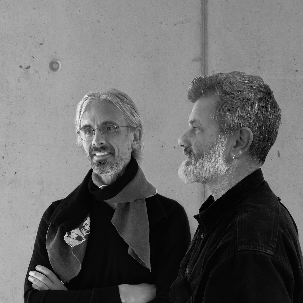 Die zwei Designer Kristof Pycke und Bruno Aeberli stehen nebeneinander vor einer betonierten Wand. Sie sind die Designer des Lampenmodells Bob von Belux.