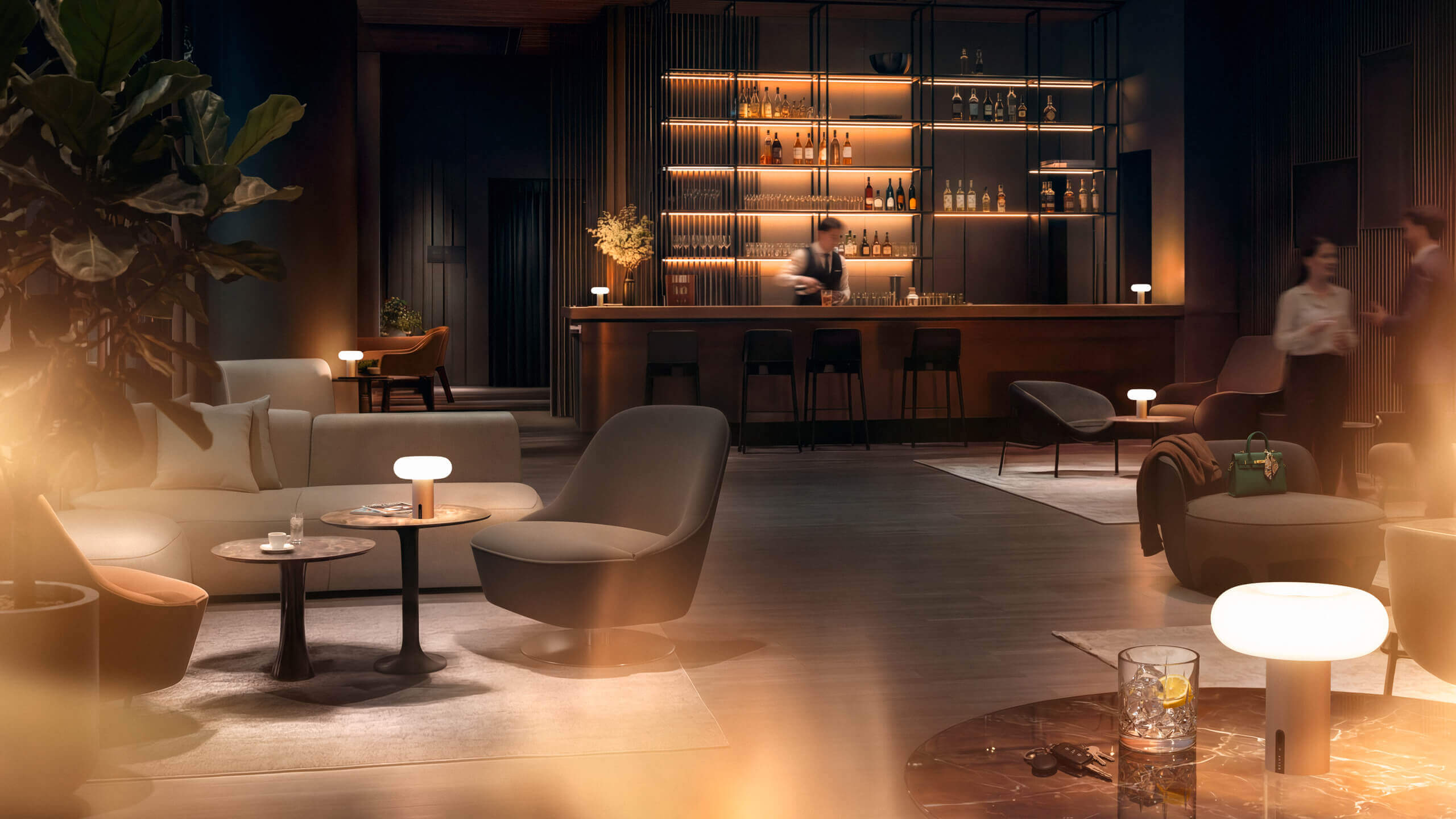 Hotel-Lounge mit Bar im Hintergrund; mehrere kleine Belux „Oto Portable“-Leuchten leuchten auf Tischen und Beistellflächen.