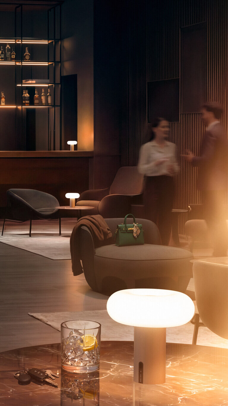 Hotel-Lounge mit Bar im Hintergrund; mehrere kleine Belux „Oto Portable“-Leuchten leuchten auf Tischen und Beistellflächen.