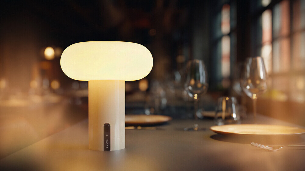 Small, bright Belux "Oto Portable" table lamp on a table, blurred glasses in the background and a warm atmosphere.