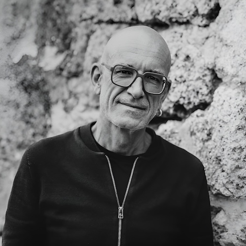 Designer Norbert Meier stands in front of a rough stone wall, smiling in a black jumper. He is the designer of the Equinox lamp model from Belux.
