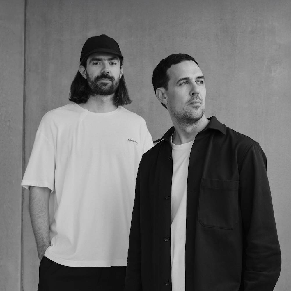 Die zwei Designer Joel Hoff und Philipp von Lintel in monochromer Kleidung posieren vor einer grauen Wand. Schwarz-weiß Portrait Foto. Sie sind die Designer der Lampenmodelle Oto Portable und Mono der Firma Belux.