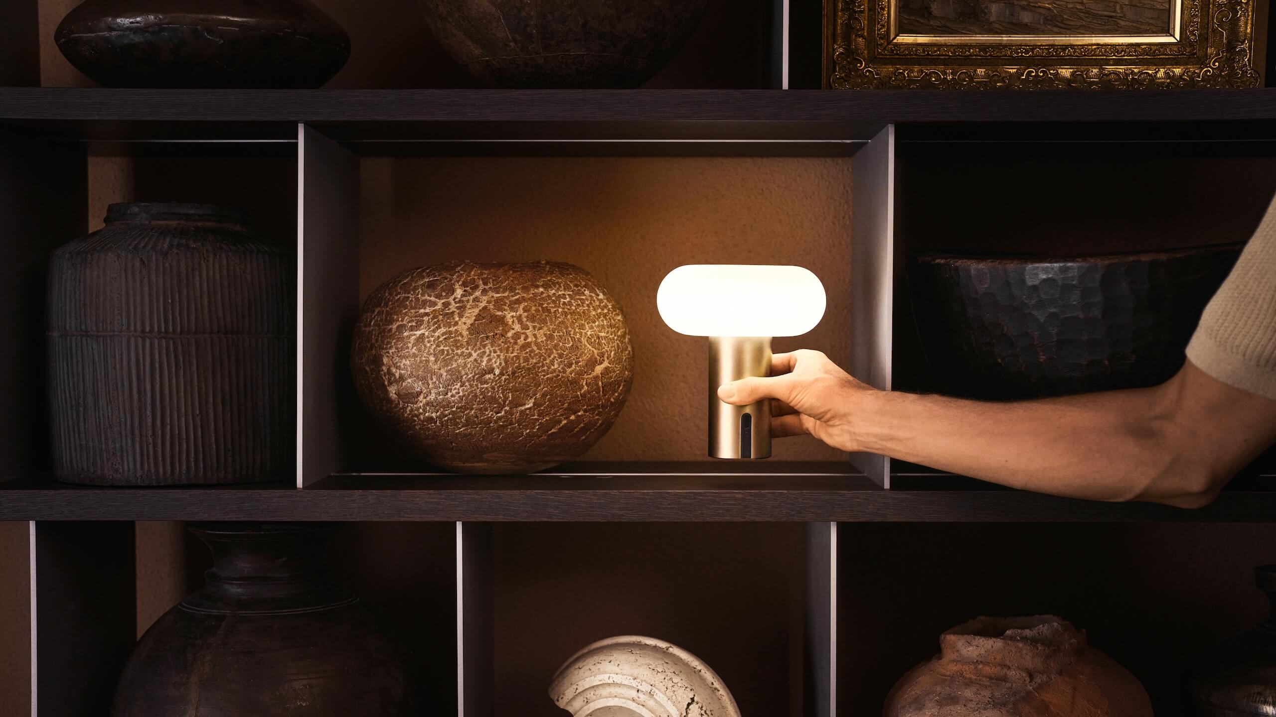 Hand places a glowing Belux "Oto Portable" on a shelf between decorative vessels.