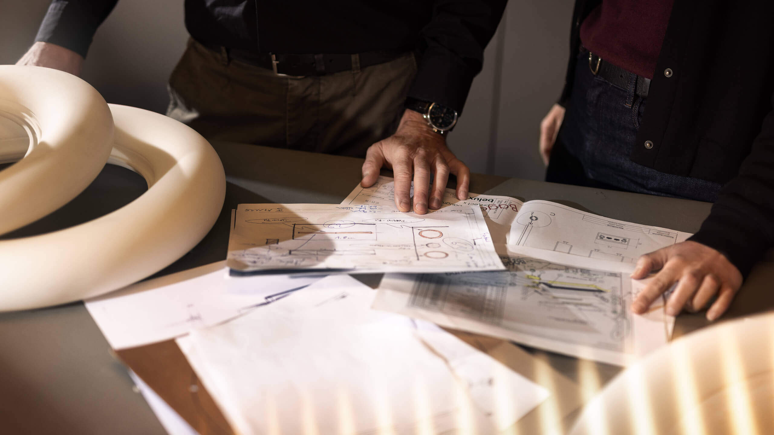 Zwei Personen betrachten technische Zeichnungen auf einem Tisch; daneben liegen helle, ringförmige Leuchtenteile.